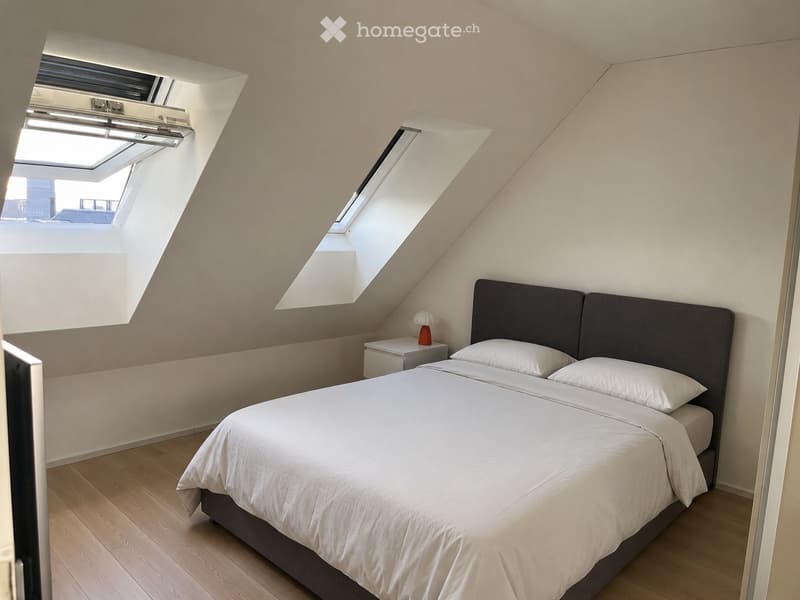 Modern bedroom with a double bed, white bedding, nightstand, and lamp. Slanted ceiling with skylights provides natural light.