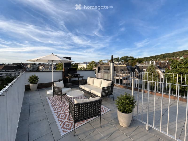 A modern rooftop terrace with a white sofa, two black wicker chairs, a small round table, a large umbrella, a grill, and potted plants, overlooking a cityscape.