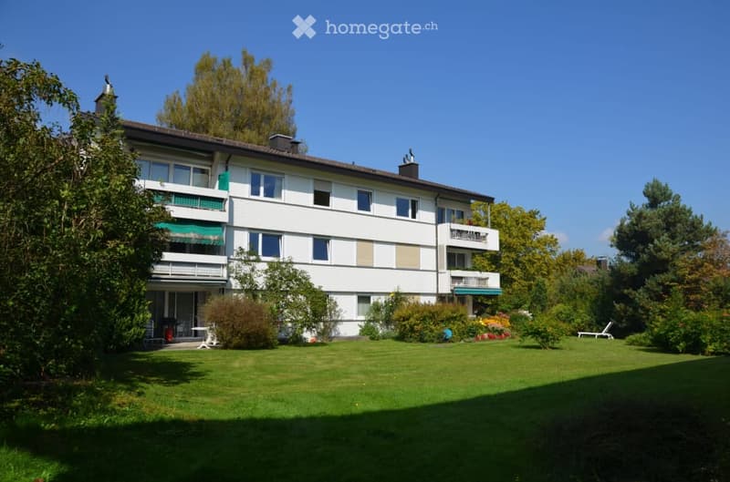 A multi-story residential building with white walls, multiple windows, balconies, and a well-maintained garden area with green lawns, trees, and outdoor furniture.