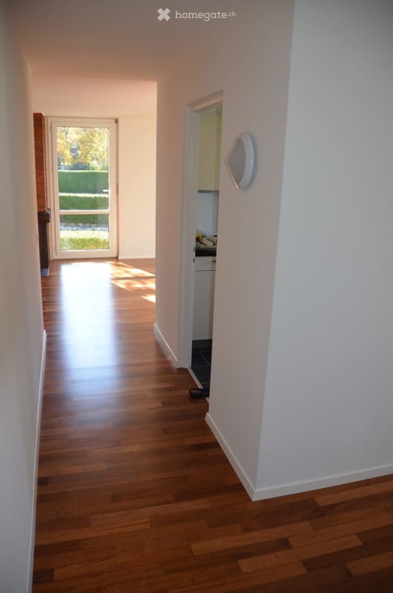 Bright hallway with wooden flooring, white walls, and a view of a kitchen area and a glass door leading to a garden.