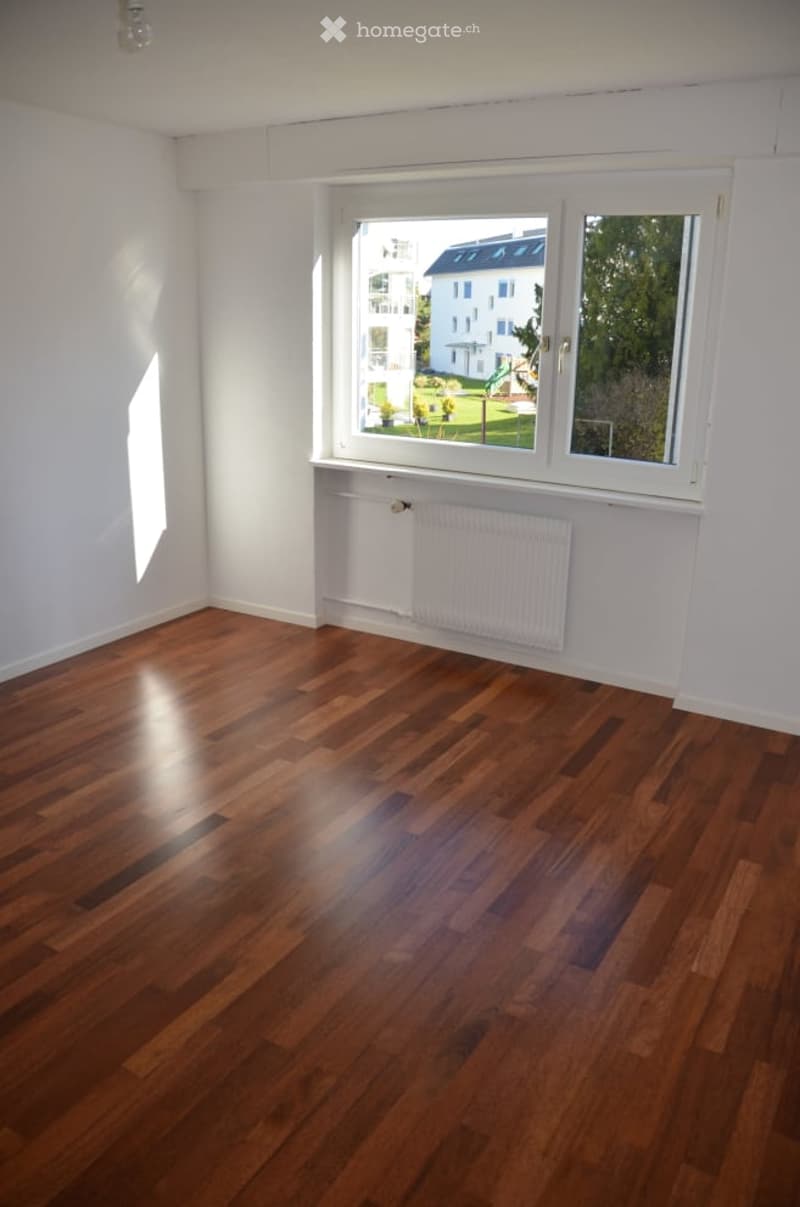 Empty room with wooden flooring, white walls, and a large window offering a view of a building and greenery outside. A radiator is visible below the window.