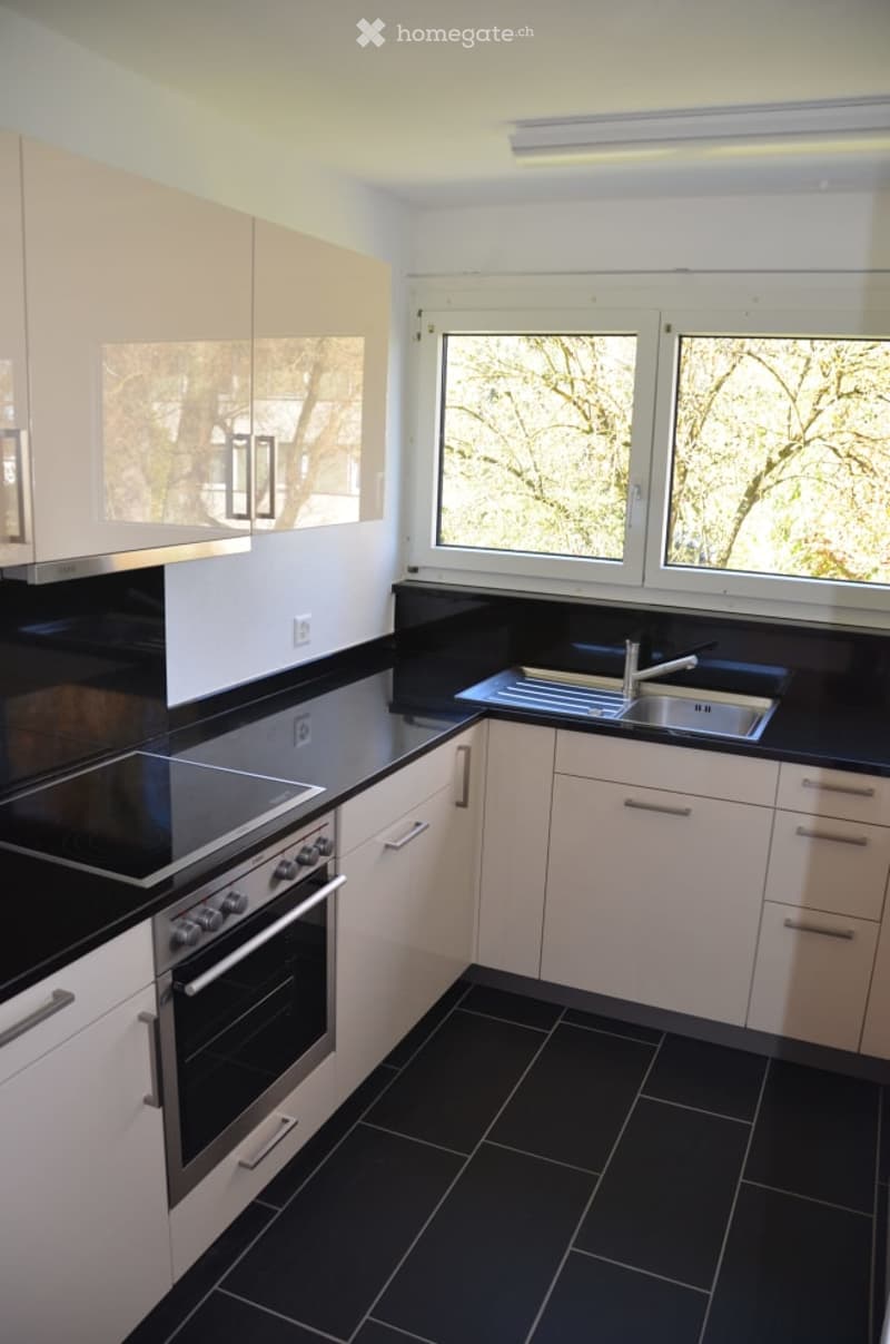 Modern kitchen with white cabinets, black countertops, and stainless steel appliances including a sink, oven, and stove. Large windows provide natural light.