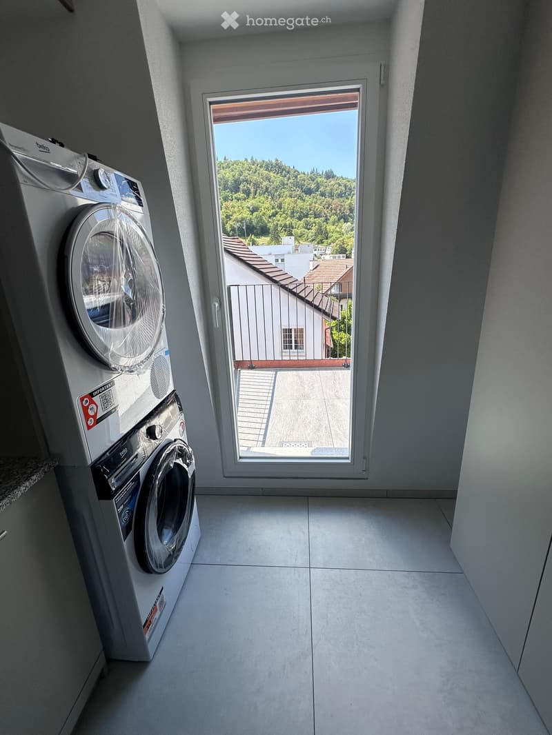 Modern laundry room with a washing machine and dryer, large window offering a view of greenery and neighboring buildings.