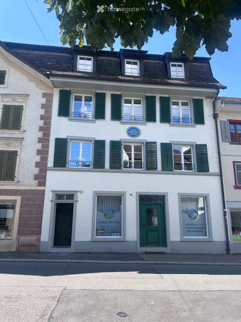Two-story building with white facade, green shutters, multiple windows, and signage for "Zurzi Spitex" and "ZVM Römischen Kaiser" on the ground floor.