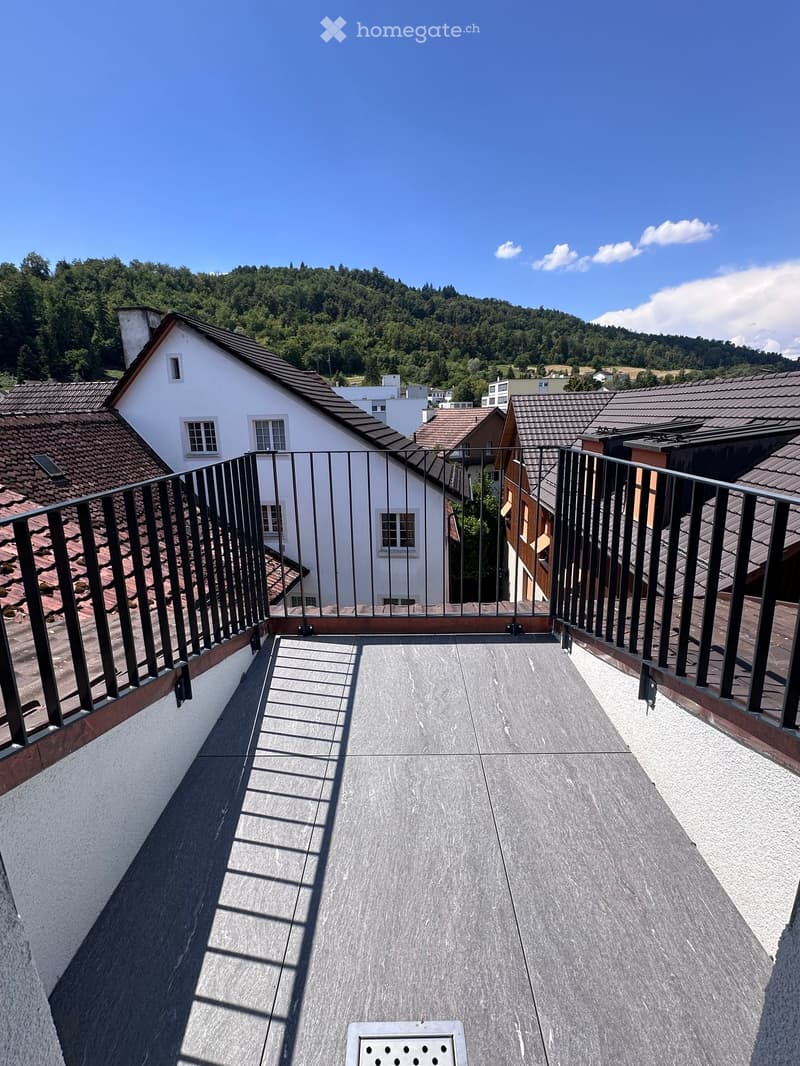 Roof terrace with grey tiled flooring, black metal railing, and scenic view of surrounding rooftops and hills.