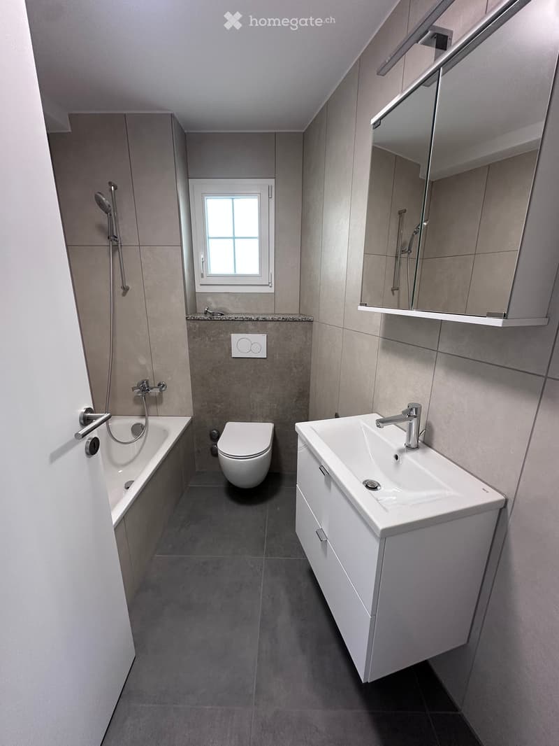 Modern bathroom with a bathtub, toilet, sink, and mirror. Features grey tiles and a window for natural light.
