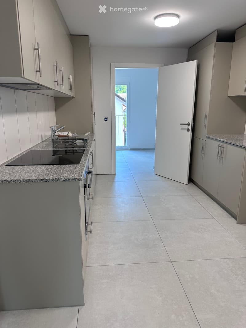 Modern kitchen with light grey cabinets, granite countertop, and a sink with a faucet. An open doorway leads to a tiled area with a view of a balcony.