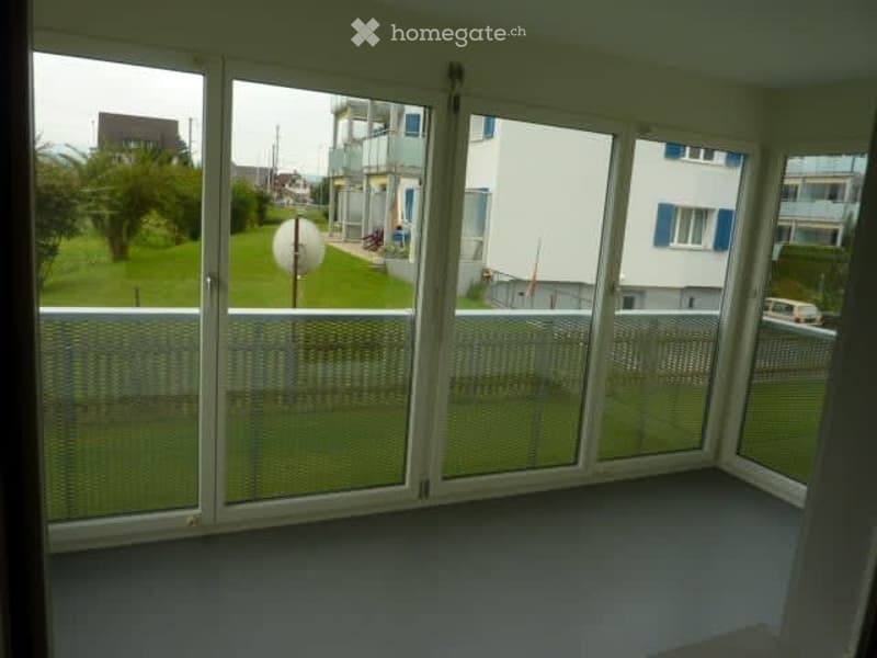 Empty room with large glass sliding doors leading to a balcony, offering a view of a green area and neighboring buildings.