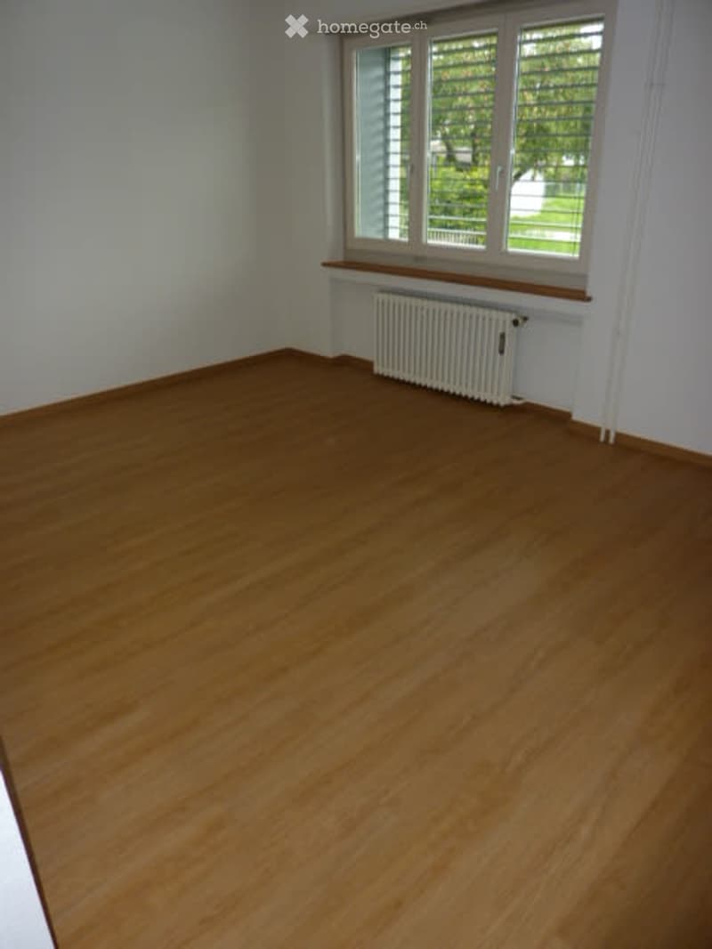 Empty room with wooden flooring, white walls, and a window with blinds. A radiator is placed below the window.