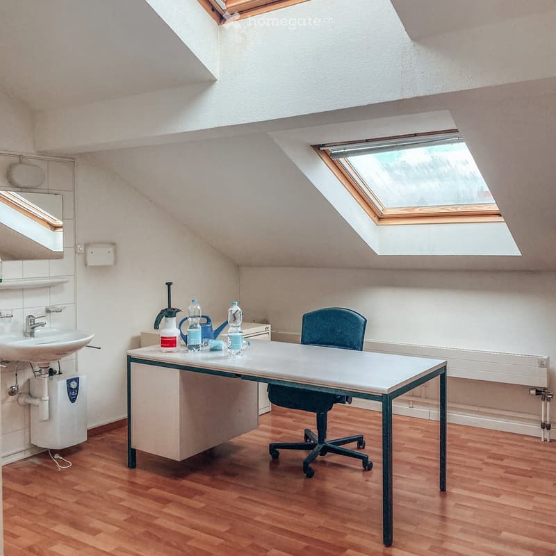 A small attic room with wooden flooring, a sink, a table with a chair, and several bottles on the table. The room has skylight windows and a radiator.