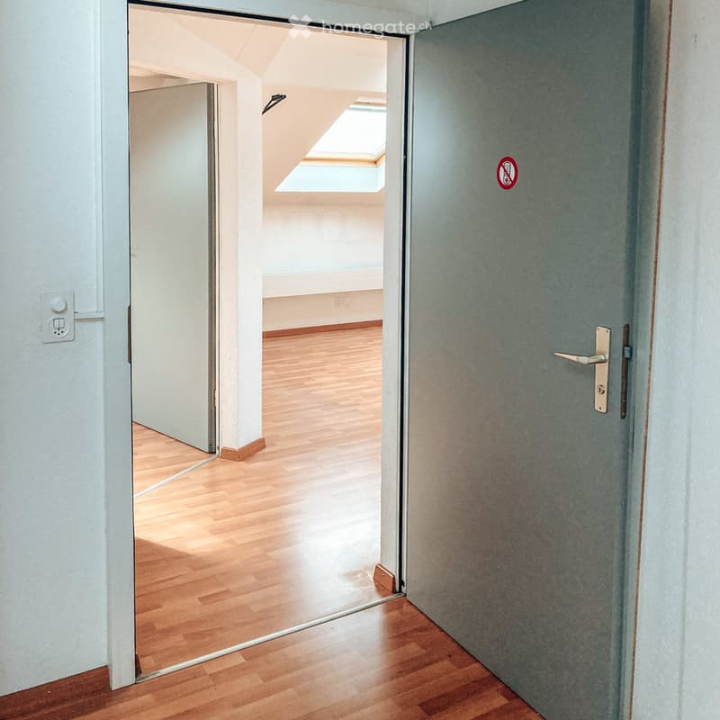 Empty hallway with wooden flooring, white walls, and a closed door on the right side. An open doorway leads to another room with a skylight window at the far end.