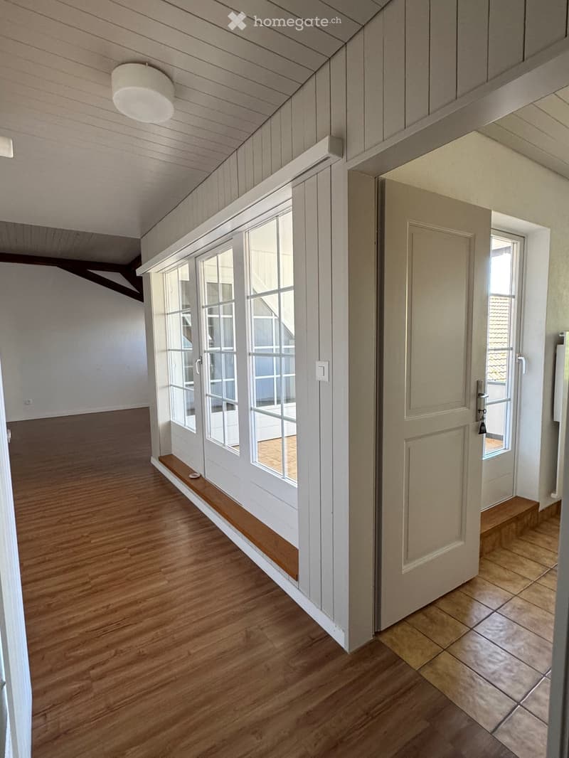 Bright hallway with wooden flooring, white walls, and large windows. Features a built-in wooden bench and a closed door leading to another room.