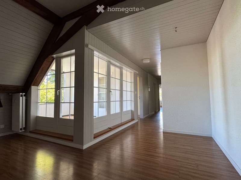 Empty attic corridor with wooden floors, white walls, large windows, and exposed wooden beams on the ceiling.