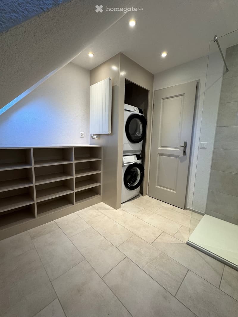 Modern bathroom with tiled floor, built-in washing machine, shelves, and a door leading to another room.