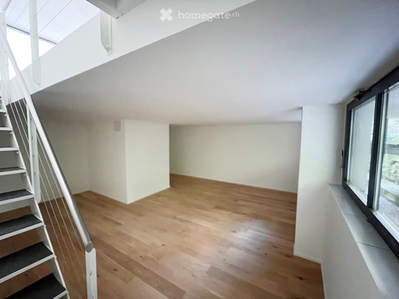 Empty room with wooden flooring, white walls, and a staircase leading upwards. Large windows on one side provide natural light.
