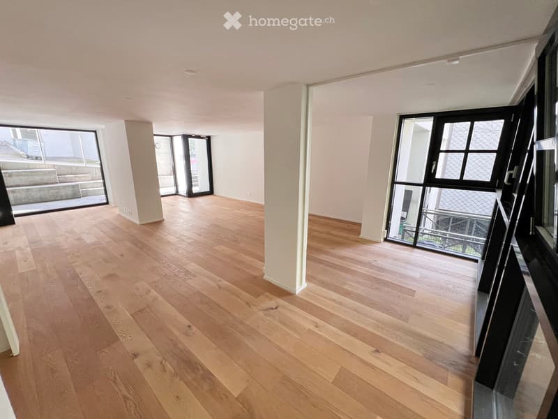 Empty room with light wooden flooring, white walls, and large windows with black frames, leading to an outdoor area.