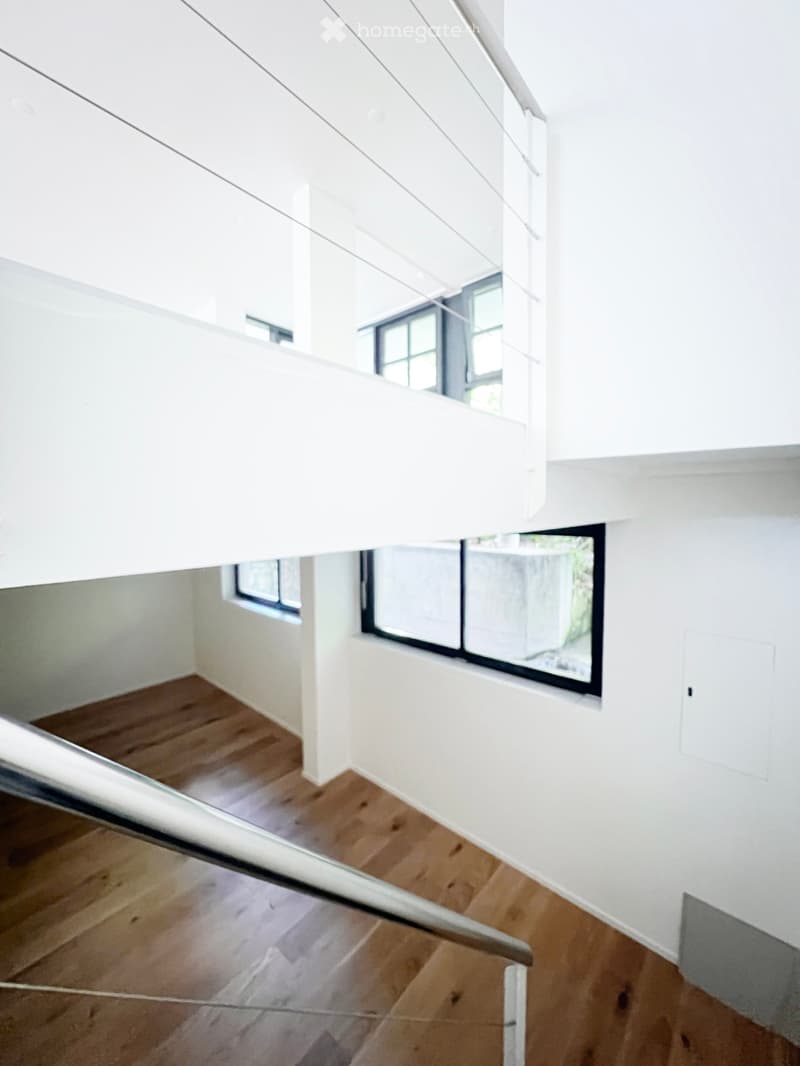 Modern, minimalist hallway with wooden flooring, white walls, and large windows allowing natural light. The staircase features a sleek metal railing and a white balustrade.