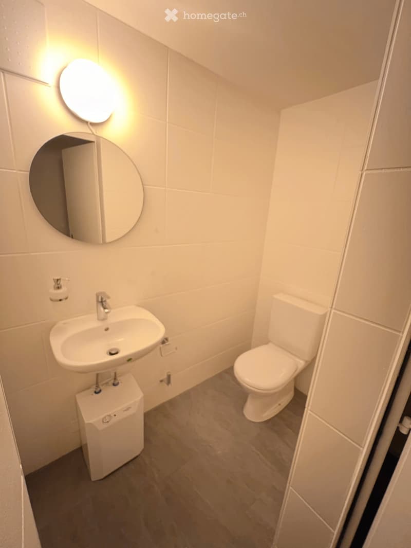 Modern bathroom with white tiles, a sink, toilet, and a round mirror above the sink.