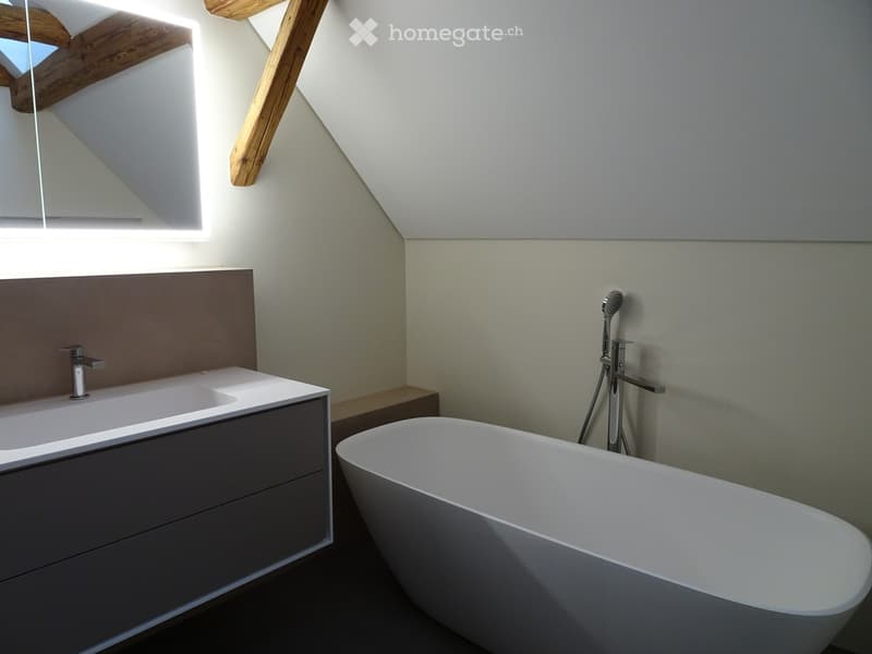 Modern bathroom with a freestanding bathtub, a sink with a sleek faucet, and a large mirror above the sink. The room features light-colored walls and wooden beams on the ceiling.