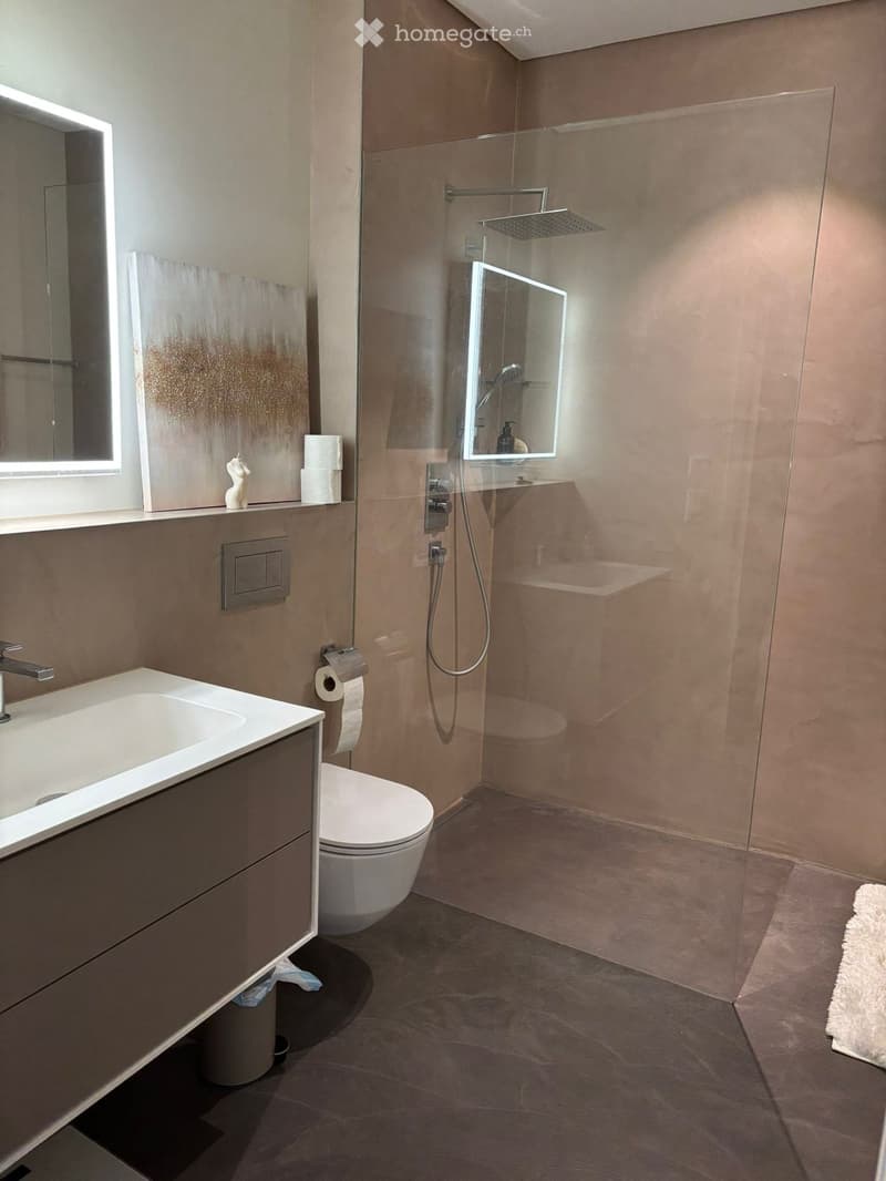 Modern bathroom with a white sink, toilet, glass-enclosed shower, and illuminated mirrors. Features beige tiles and dark flooring.