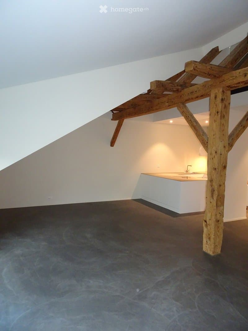 Open loft space with exposed wooden beams and a polished concrete floor. A small white kitchen area is visible in the background, but the main room is completely empty with no furniture or equipment.