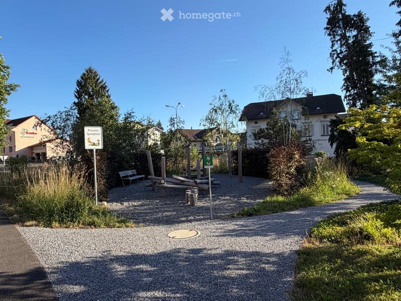 Private playground area with gravel paths, wooden play structures, benches, and surrounding greenery.