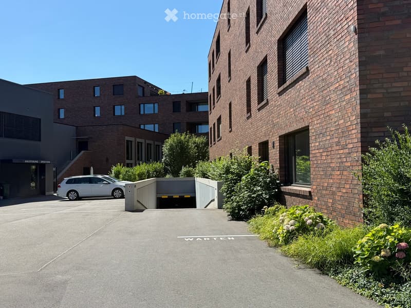 Modern multi-story brick building with large windows, a raised ground floor, and a parking area with a white car.