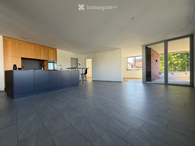 Modern kitchen with grey and wooden cabinetry, grey countertop, grey tiled floor, and a small dining area with a table and stool. Large windows and sliding glass doors lead to a balcony.