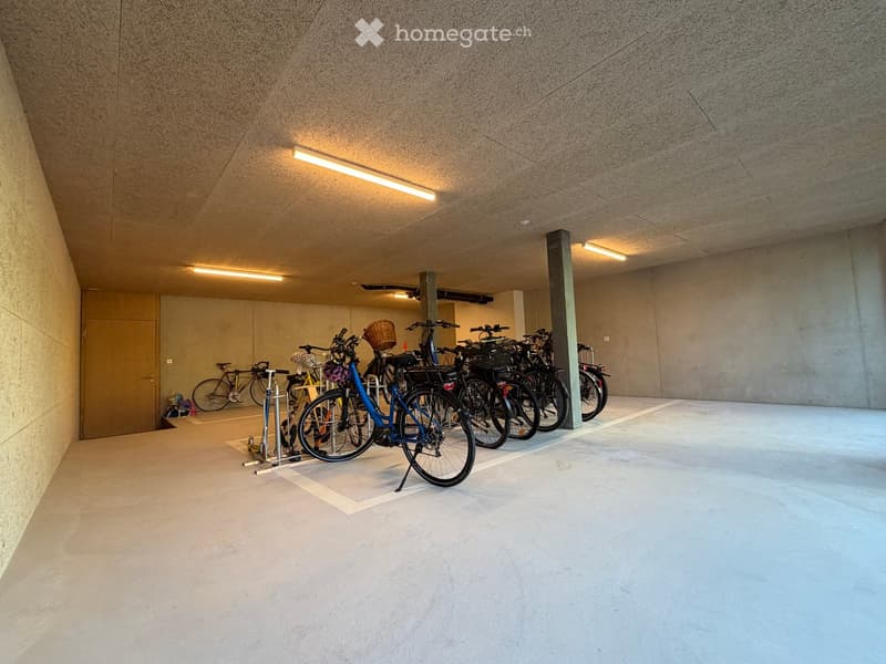 Modern indoor bicycle parking area with concrete walls and ceiling, several bicycles parked on racks, and a door on the left side.