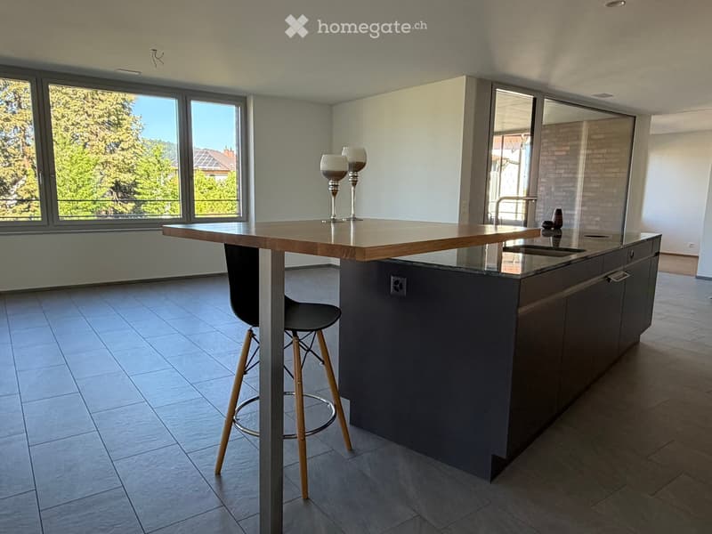 Modern kitchen with a central island, dark cabinets, and a light wooden countertop. Two stools are placed under the island, and there is a sink with a modern faucet. The space is well-lit with large windows offering a view of greenery outside.