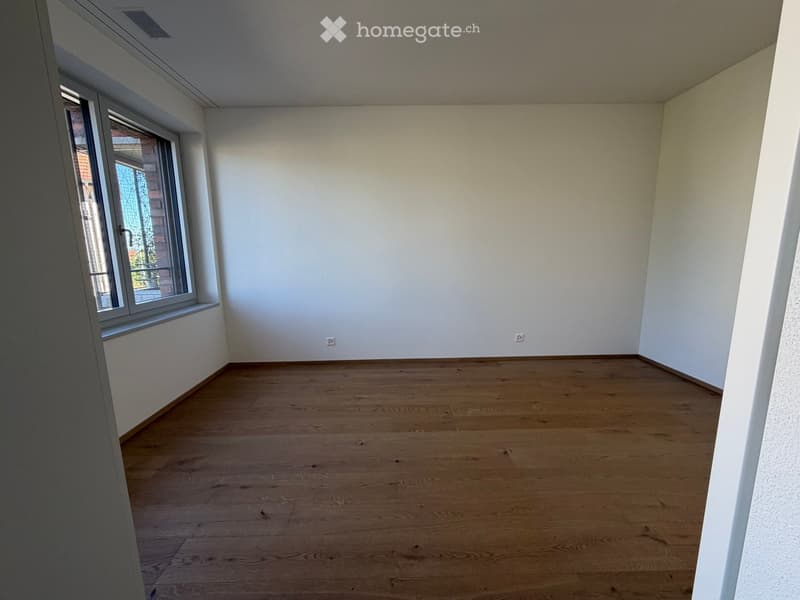 Empty room with wooden flooring, white walls, and a large window with a view of a brick building outside.