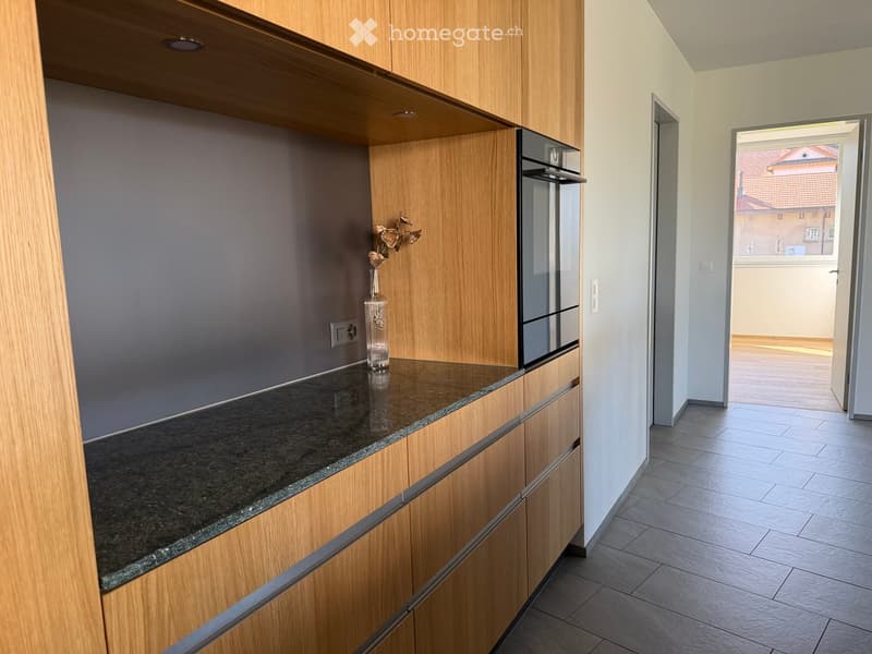 Modern kitchen with wooden cabinets, granite countertop, and a vase with flowers on the counter. The hallway leads to another room with tiled flooring.