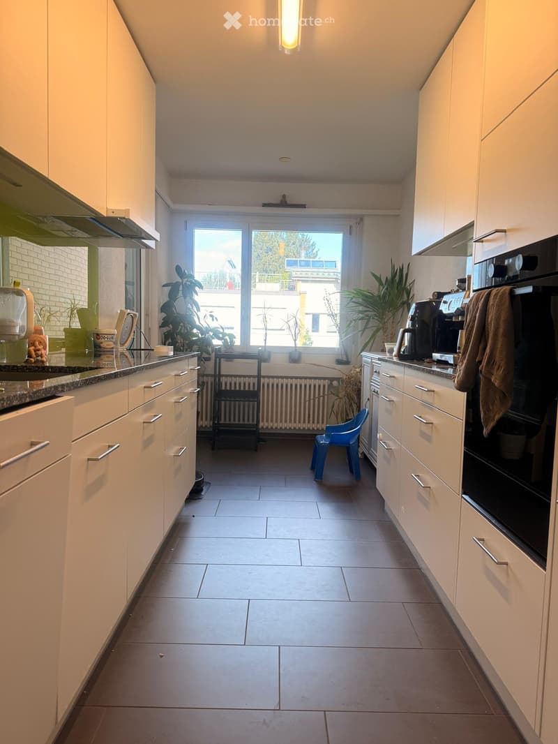 Modern kitchen with white cabinets, granite countertops, and a large window overlooking a balcony. Appliances include a sink, fridge, oven, and coffee maker. A small blue stool is placed near the window, and potted plants are on the windowsill.