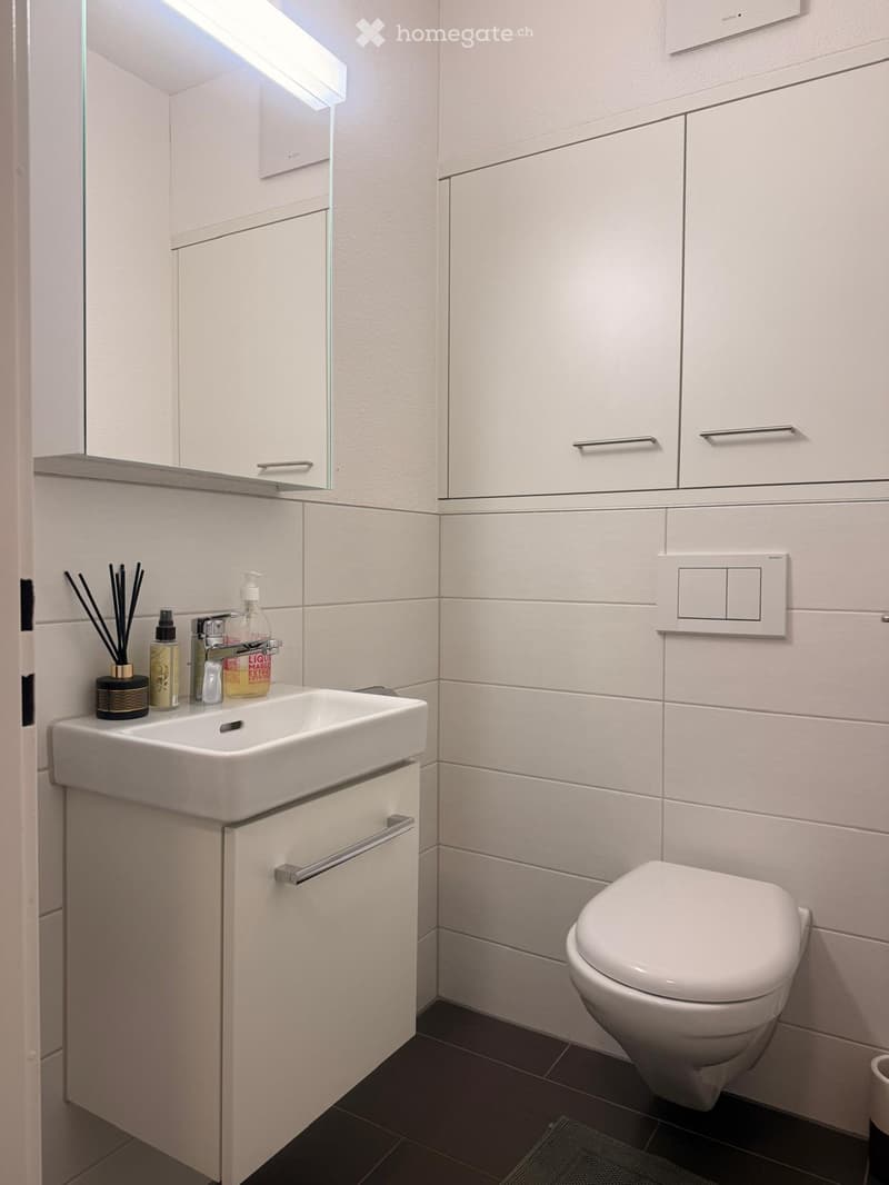 Modern bathroom with white tiles, a sink with a faucet, a toilet, a mirrored cabinet, and a light switch on the wall.