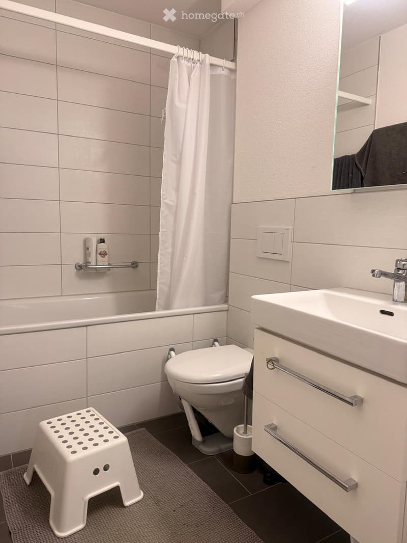 Modern bathroom with white tiles, bathtub, toilet, sink, mirror, and a small white stool. Includes a shower curtain and a bottle on the shelf.