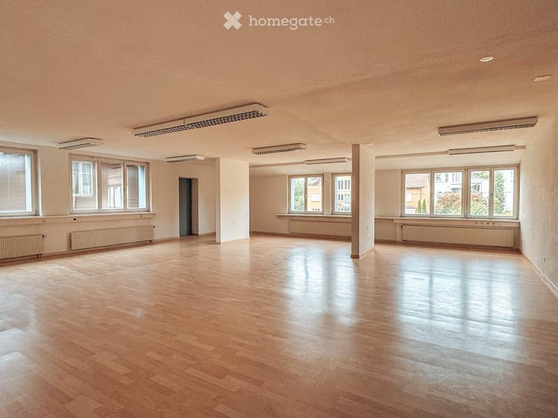 Empty room with wooden flooring, large windows, radiators, and a central column. No furniture or equipment present.