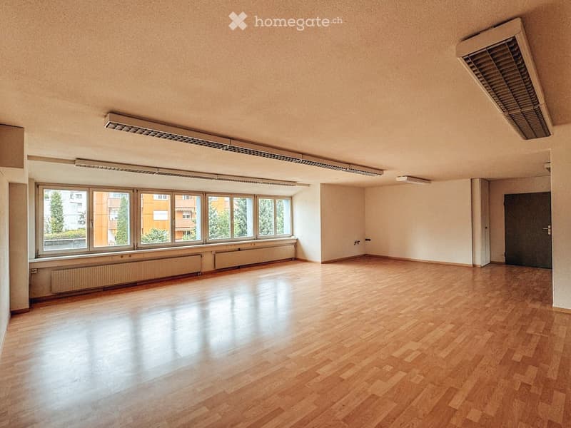 Empty room with wooden flooring, large windows, radiators, and a door. No furniture or equipment present.