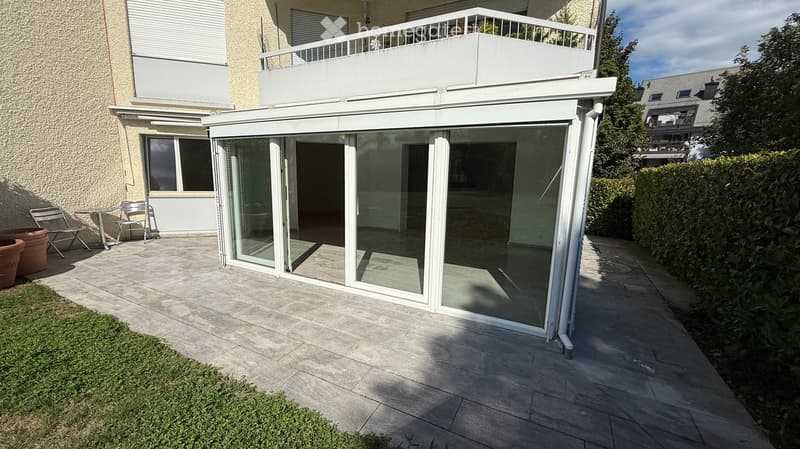 Outdoor patio area with a glass enclosure, tiled flooring, and a small seating area with chairs and a table. The patio is adjacent to a building with a balcony above.
