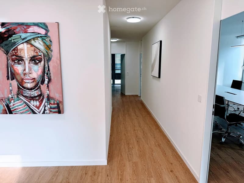 Modern hallway with light wooden flooring, white walls, and recessed ceiling lighting. Features a large abstract painting on one wall and a glimpse of an office area with a desk and chair at the end.