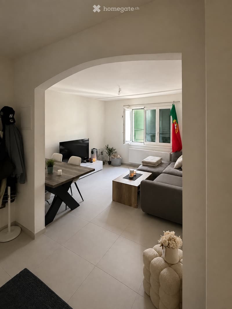 Modern living room with white walls, light-colored tiles, and a high ceiling. Features a grey sectional sofa, wooden coffee table, dining table with chairs, and a television on a stand. A small plant and decorative items are placed around the room.