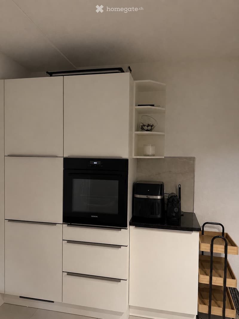 Modern kitchen with white cabinets, a built-in oven, a coffee maker, and a small wooden cart with shelves.