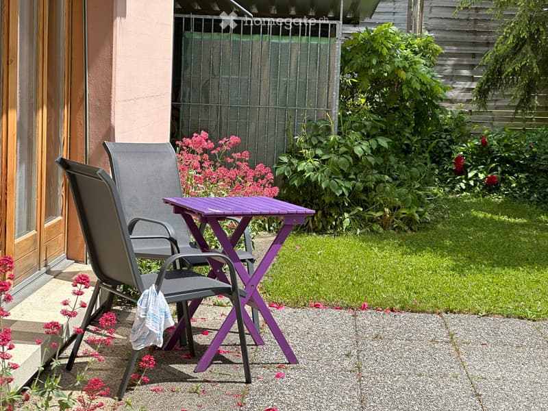 Outdoor patio area with a purple folding table and two black folding chairs, surrounded by greenery and flowers.