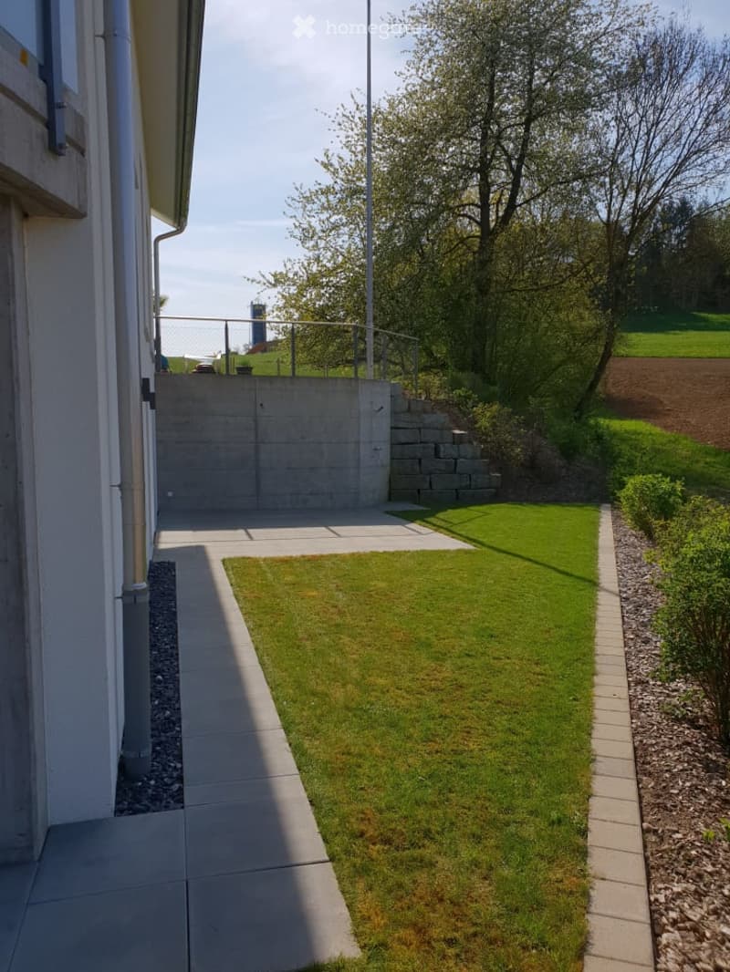 Well-maintained garden area with a concrete pathway, neatly trimmed grass, gravel borders, and a small concrete retaining wall. The area is bordered by a building on one side and a fence on the other, with trees and open space in the background.