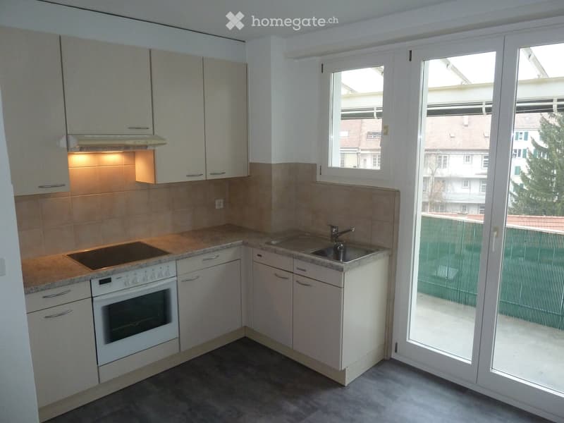 Modern kitchen with white cabinets, granite countertops, built-in oven, and a sink with a faucet. Large window and sliding glass door lead to a balcony.