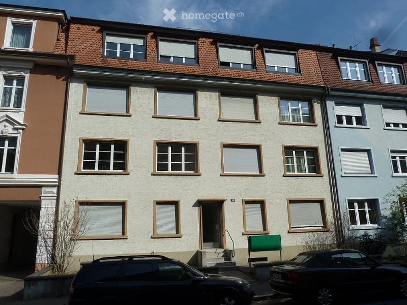 Multi-story residential building with beige facade, multiple windows with shutters, and a small entrance staircase leading to the main door.