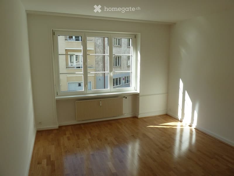 Empty room with wooden flooring, white walls, and large windows showing a view of neighboring buildings.