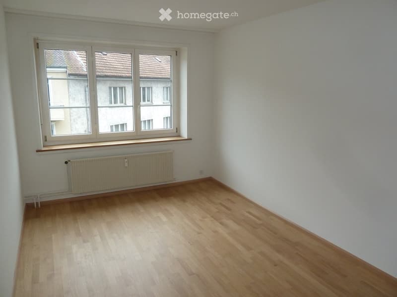 Empty room with light wooden flooring, white walls, and a large window with a radiator below it.