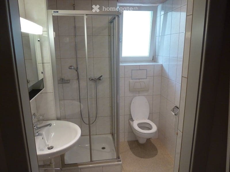 Modern bathroom with a white tiled shower stall, a white toilet, and a white sink with a chrome faucet. The walls are tiled in light beige, and there is a small window above the toilet.