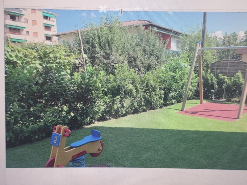 A well-maintained garden area with artificial grass, a wooden rocking horse, and a swing set in the background, surrounded by lush greenery and residential buildings.