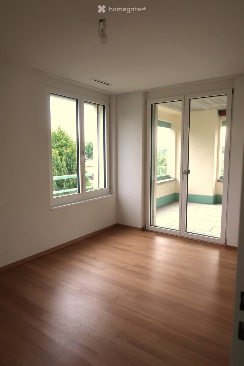 Empty room with wooden flooring, white walls, large windows, and a sliding glass door leading to a balcony.